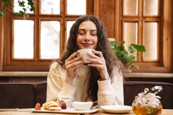Mulher jovem com cabelos ondulados, sorrindo com os olhos fechados, segurando uma xícara branca de chá perto do rosto em um ambiente rústico e acolhedor, com um bule de vidro e petiscos sobre a mesa. Representa bem-estar e prazer.